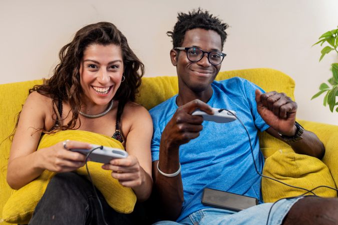 A joyful diverse couple laughing at the camera while playing video games together on a sofa at home.