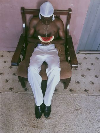 Topless man in light pants holding a slice of watermelon sitting wooden armchair