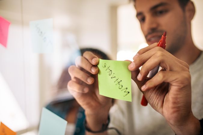 Man writing on sticky notes with marker pen and pasting on glass board in office
