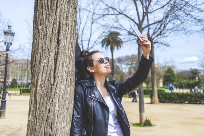 Side view of a young laughing hipster woman standing and leaning on a tree at park in sunny day while using a mobile phone to take a selfie