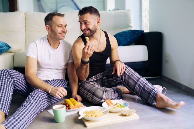 Happy male couple enjoying meal at home sitting on floor