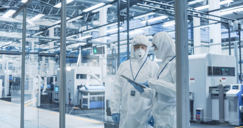 Industrial Scientists Wearing Disposable Protective Suits, Masks, and Goggles at a Production Plant. Specialist Using Tablet, Analyzing Potential Work Hazards and Contamination at a Workplace