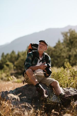 Mature man on hiking trip sitting on a rock with a digital camera