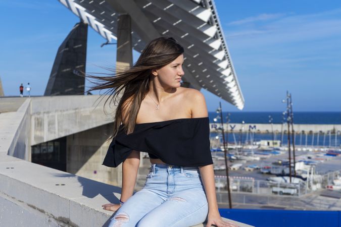 Young woman with long hair sitting on promenade against harbor while looking away