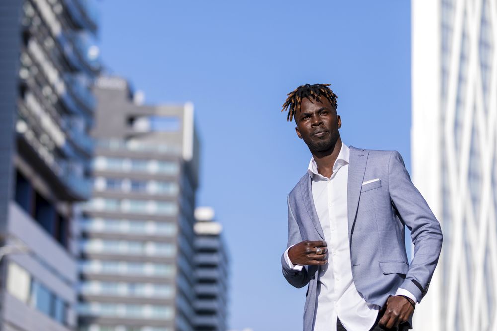 Front view of a Black man wearing elegant clothes standing in the street while looking in the distance in sunny day