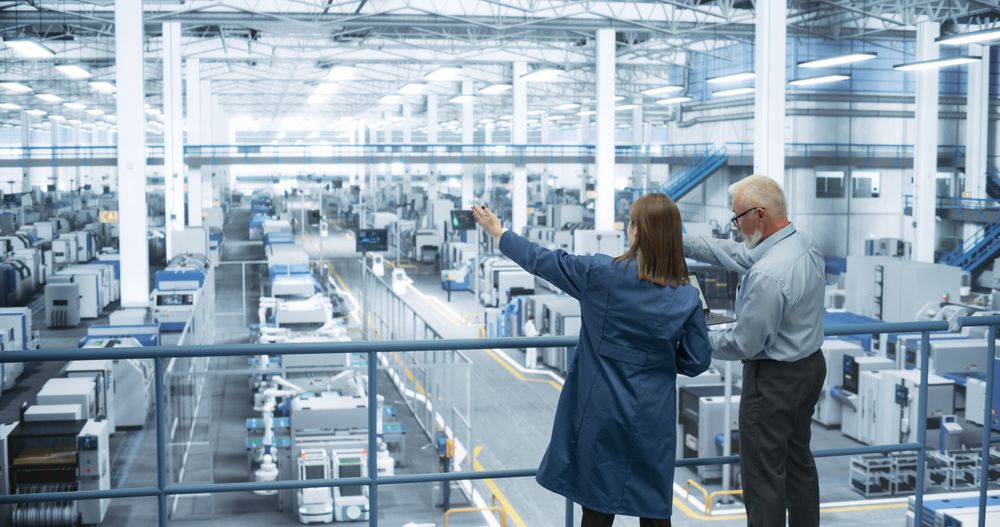 Male Engineer and Female Project Manager Standing on a Platform, Talking, Using Laptop Computer and Monitoring Production at a Modern Automated Electronics Manufacture with AI Robotic Arms