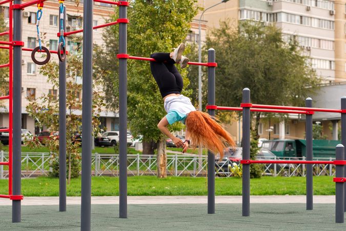 Woman performing acrobatic move on outdoor horizontal bar in park.