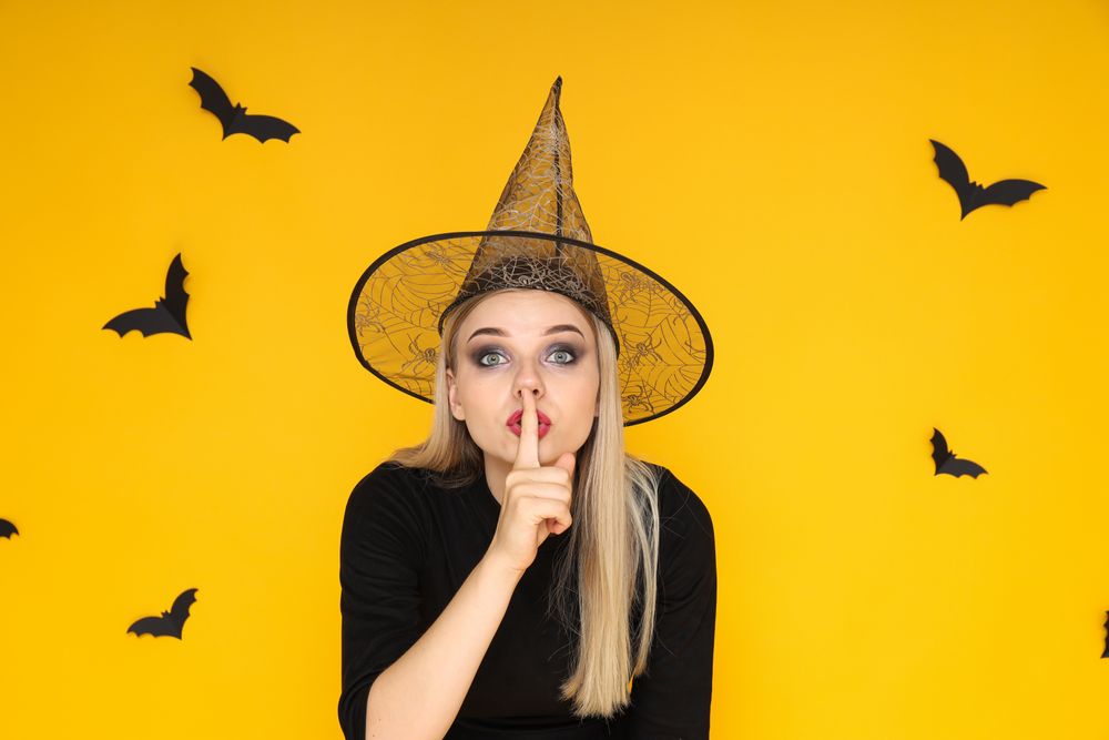 A woman in a Halloween costume shows "shh", a silent gesture, on a yellow background