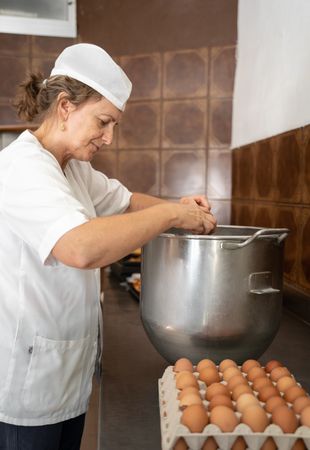 Baker woman adding eggs to a big industrial pan to prepare cupcakes
