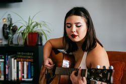 Young Asian woman sitting on orange chair playing guitar with bookshelf in background