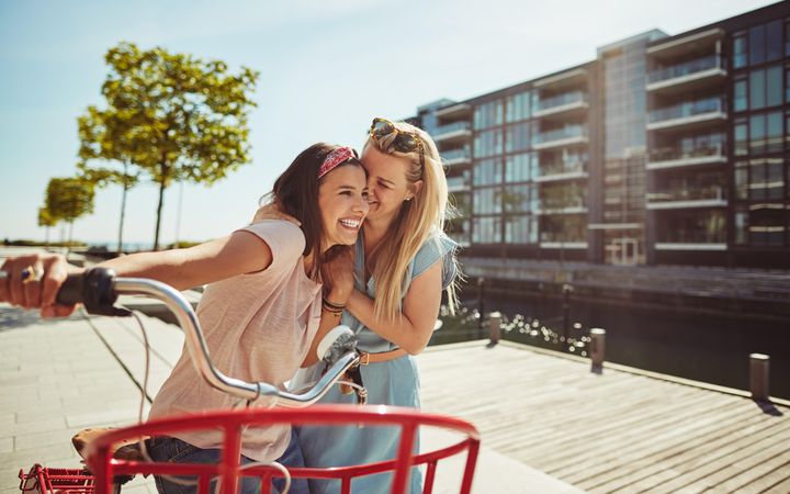 Two close female friends laughing while walking along a riverwalk with bike on sunny day, copy space