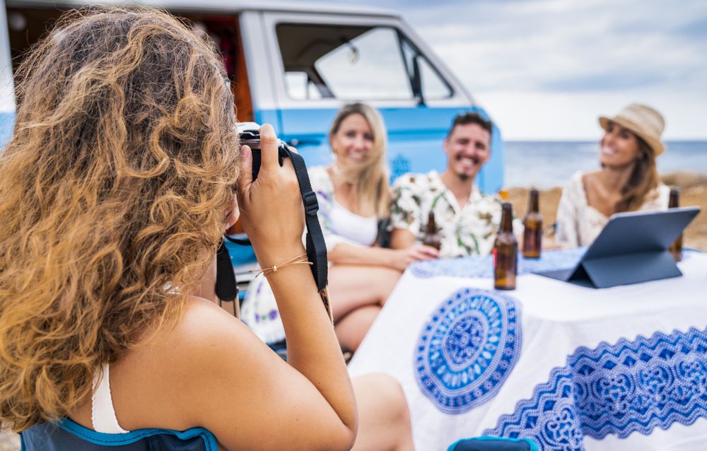 Joyful company sitting at table with drinks on beach while friend taking photo