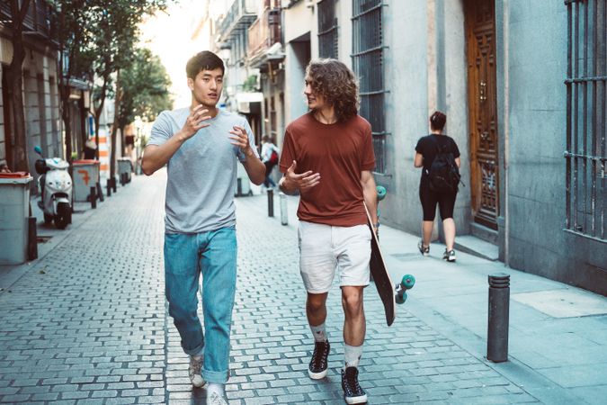 Cool multiracial male friends chatting while walking on street with longboard