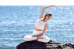 Young woman stretching her side in lotus pose on rocky beach