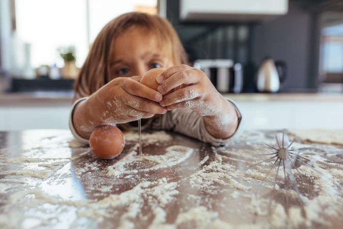 Little girl breaking an egg on kitchen counter covered with flour