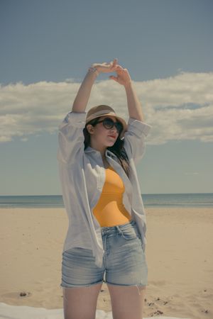 Woman in denim shorts wearing hat and sunglasses raising both arms and standing on beach