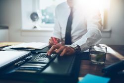 Man in shirt and tie sitting at desk with calculator in sunny office