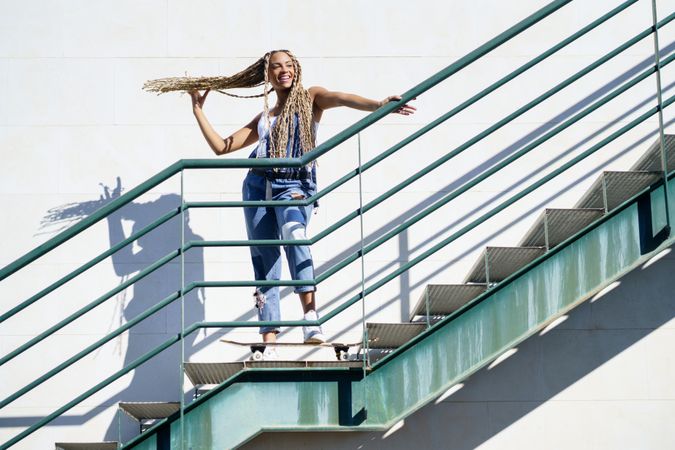 Female in denim overalls standing on skateboard on stairs playing with her hair