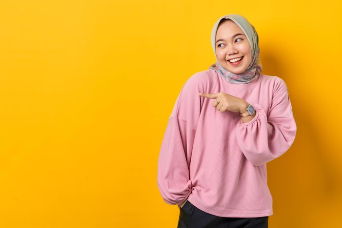Shy and happy Muslim woman in scarf and pink sweater gesturing towards copy space studio shoot