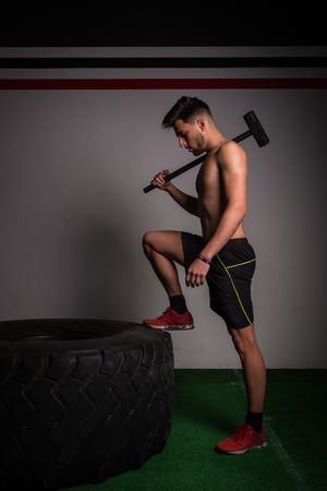 Shirtless guy concentrated in sportswear with hammer hitting on big tire in gym