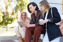 Female friends sitting on stairs together outside with coffee and digital tablet