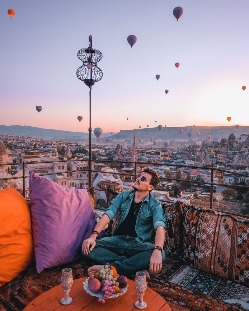 Man sitting on cushion on roof of building under sky filled with hot air balloon
