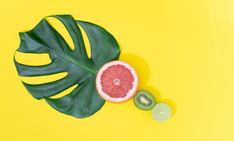 Monstera leaf with red grapefruit, lime and kiwi on bright yellow background