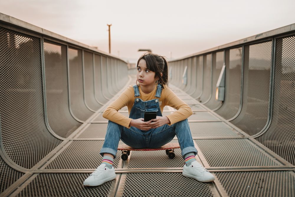 Girl with smartphone sitting on skateboard on walkway