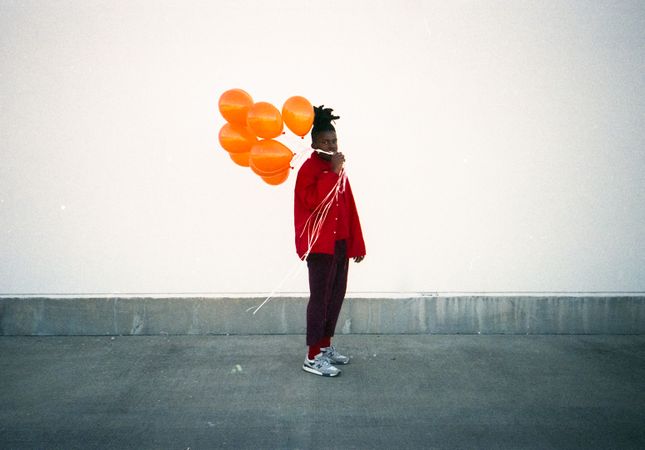 Man in red shirt holding orange balloons standing against light wall