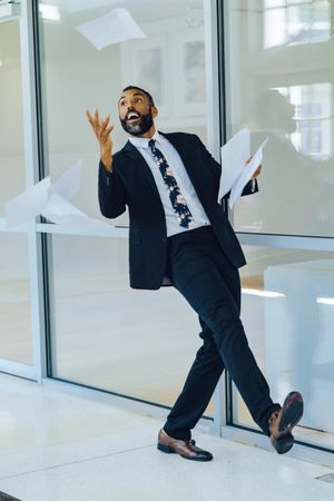 Smiling businessman in suit and tie throwing papers in the air in office