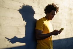 Man checking his phone in the sun against a wall, with shadow