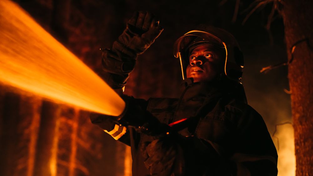 Black Fireman Holds High-Pressure Water Hose, Battling Bushfire.