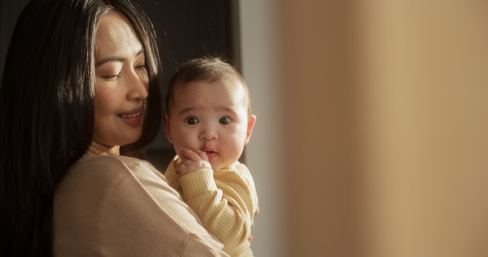 Portrait of a Young Asian Woman Holding her Baby in her Arms While Standing Next to a Window at Home.