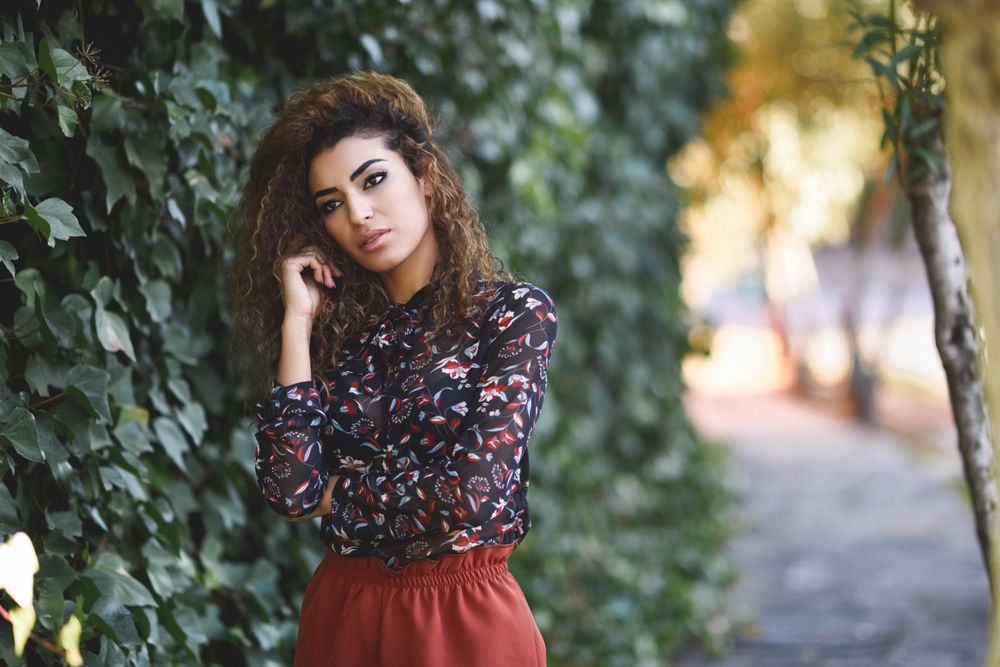 Arab woman wearing casual clothes in the street lined with greenery with hand to cheek