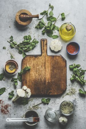 Empty wooden cutting board on grey concrete kitchen table with herbs ,spices, garlic,oil and mortar and pestle.
