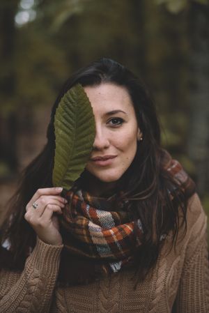 Side view of young woman in hat wrapping in checkered scarf while standing on dry leaves in autumn forest looking up