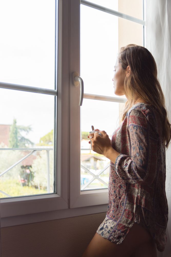woman in a shirt with a cup of coffee or tea looking out the window in the morning