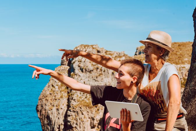 Stylish woman with tablet in hands and mature woman pointing with finger to blue sea