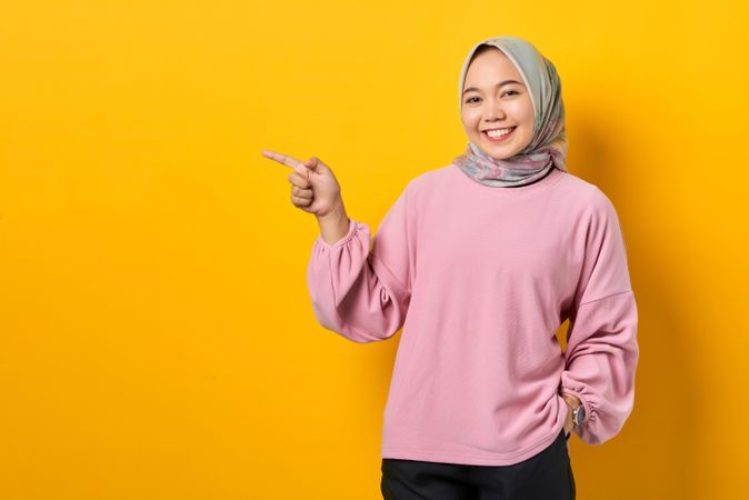 Happy Muslim woman in scarf and pink sweater pointing towards copy space studio shoot