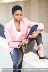 Female wearing suit with pink jacket sitting on ledge checking smartphone bxpYrb