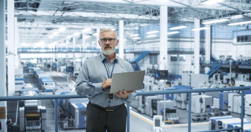 Portrait of a Handsome Mature Engineer with a Beard and Glasses Using Laptop Computer and Looking at Camera. Male Specialist Working in a Modern Electronics Factory