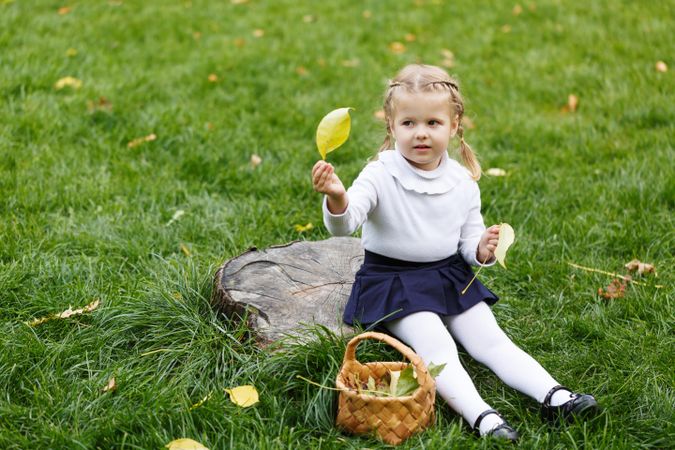 Young blonde girl sitting in the grass collecting leaves with basket