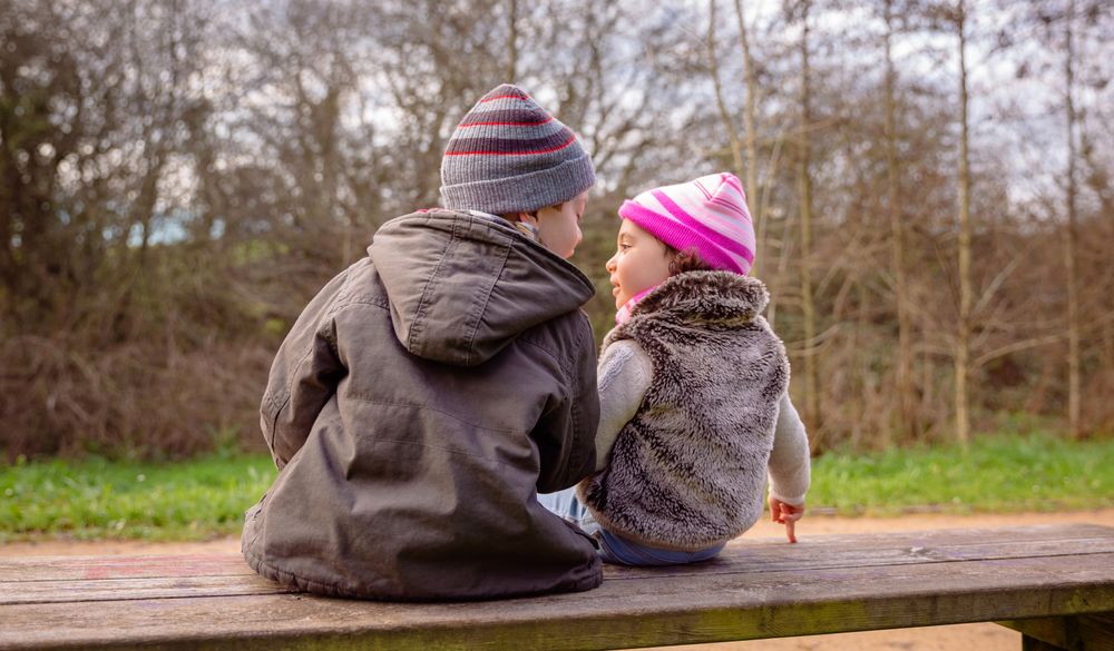 Back of little boy and girl sitting on park bench talking in forest