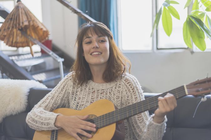 Female strumming guitar in living room of bright loft with plants in background