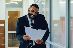 Happy businessman in suit and tie with papers walking in office while speaking on smart phone