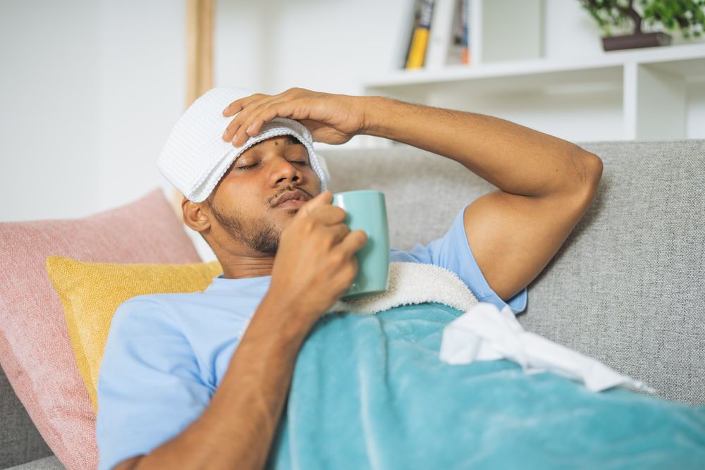 Young man with fever, drinking a cup of tea, lying down on the couch