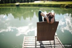 Back view of woman sitting on wooden lounge chair on a dock by lake 0L2xP4