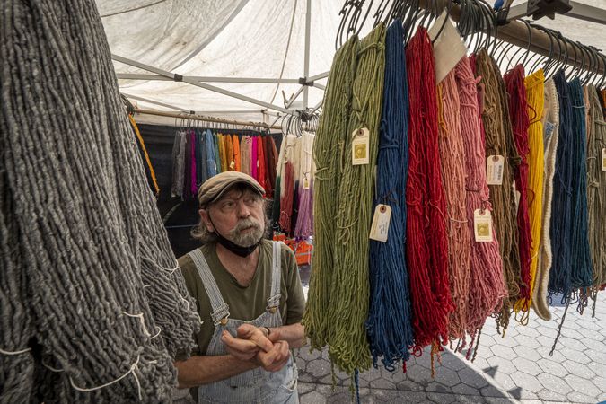 Man with overalls and cap stands near brightly colored sheep wool at a farmers market