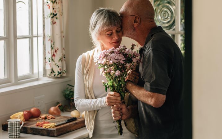 Older couple standing in kitchen holding a bunch of flowers