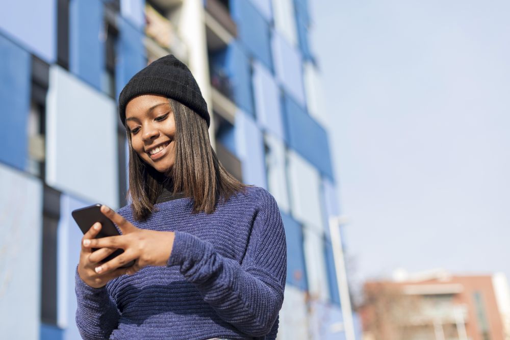 Female in hat and blue sweater checking cell phone outside blue building